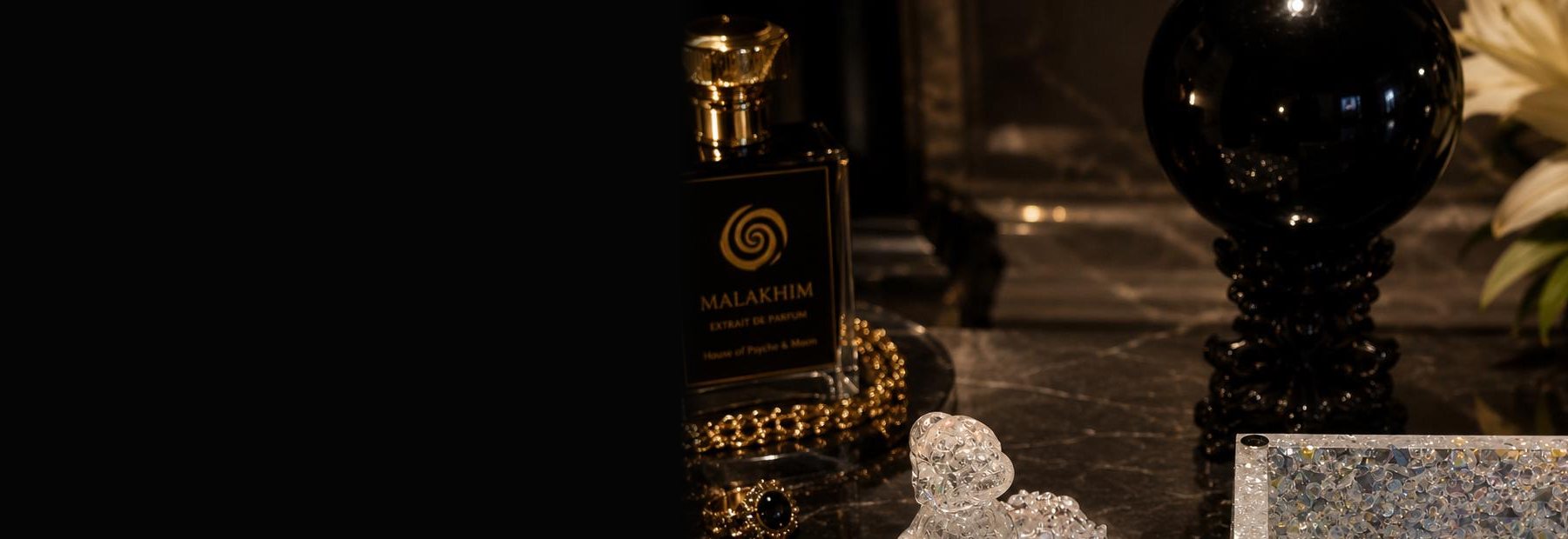 A bottle of Malakhim extrait de parfum by House of Psyche & Moon on dark marble, beside a black sphere, white lilies and candlelight.