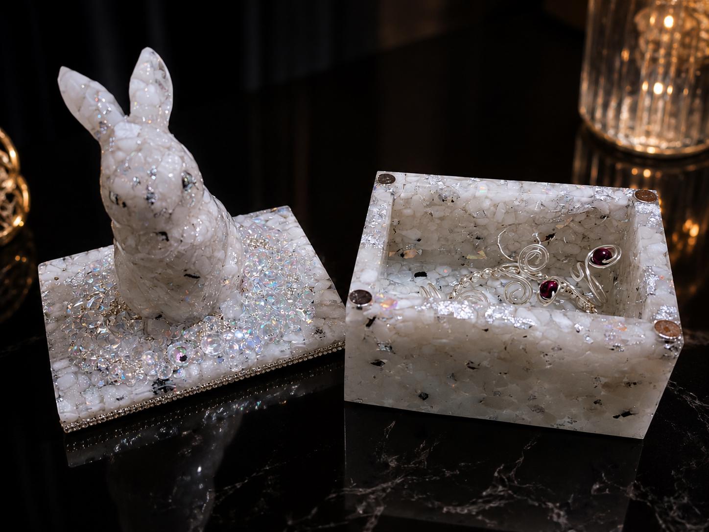 The Moonstone Hare box open beside its lid, the sculpted white hare seated on top, pale stone and silver leaf catching candlelight against black.